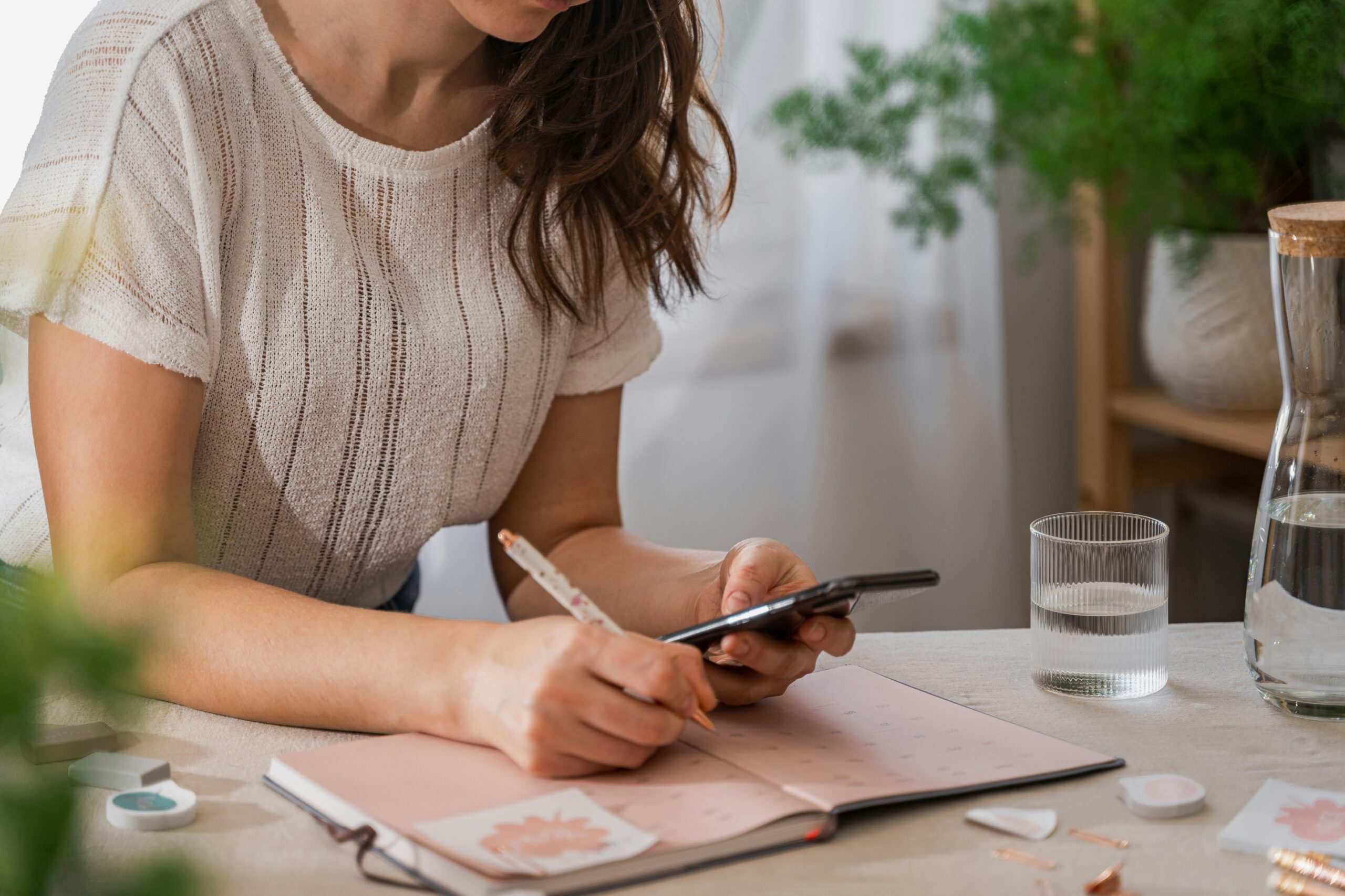 Donna che scrive su un'agenda rosa con una penna mentre consulta il telefono — riflessione e autovalutazione prima di intraprendere una nuova professione
